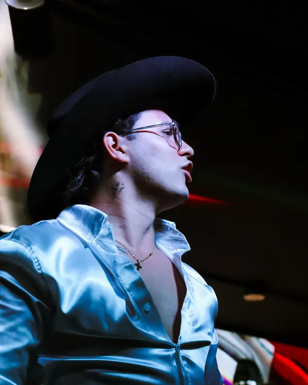 Ricardo de perfil con sombrero negro y camisa satinada, bajo luces rojas